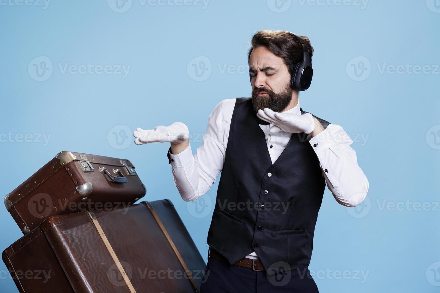 Silly hotel porter listens to music on camera, having fun with songs and dance moves. Skilled elegant doorkeeper feeling funky and using headphones, works in hospitality industry. photo