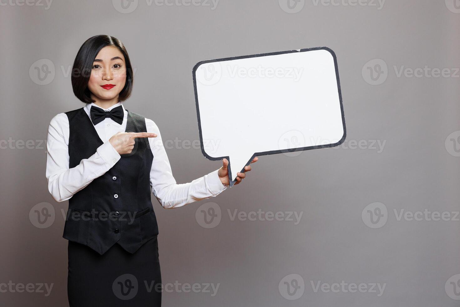 Attractive asian woman receptionist pointing at white blank speech bubble and looking at camera. Restaurant employee in waitress uniform holding dialog balloon banner mockup portrait photo