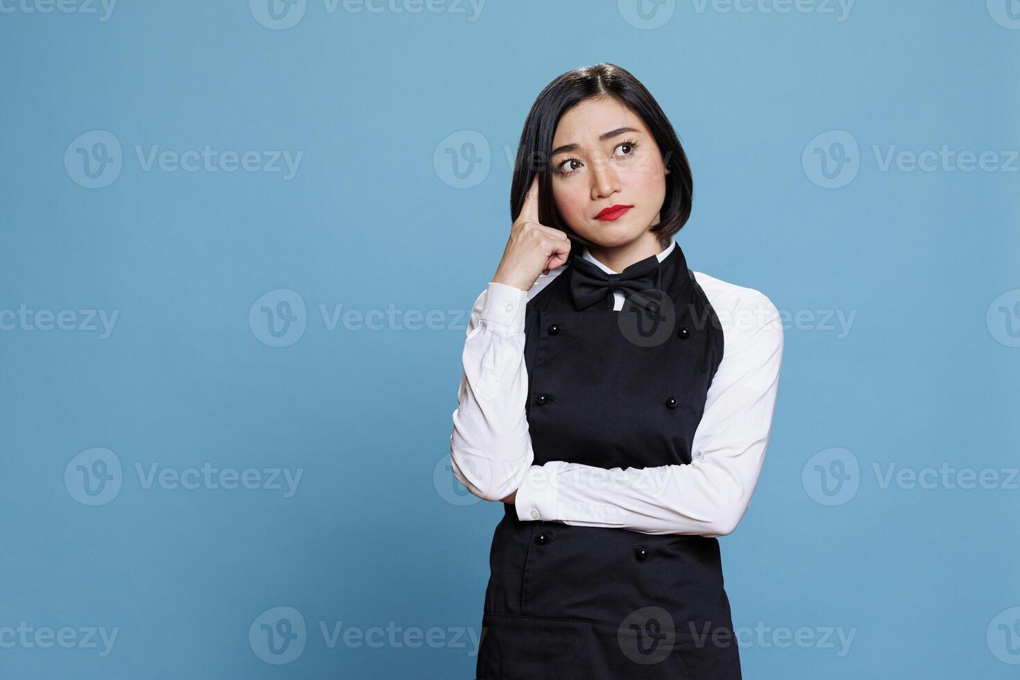 Young asian waitress with pensive facial expression holding finger on head and looking away. Attractive woman receptionist wearing uniform deep in thought posing in studio photo