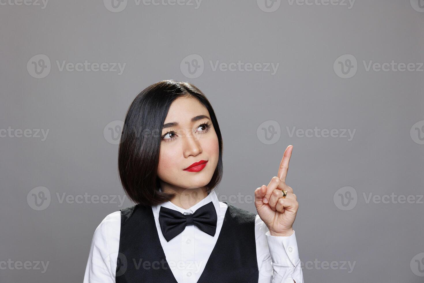Young attractive asian woman receptionist pointing and looking upwards, showcasing direction. Restaurant waitress wearing uniform showing with finger up while posing in studio photo