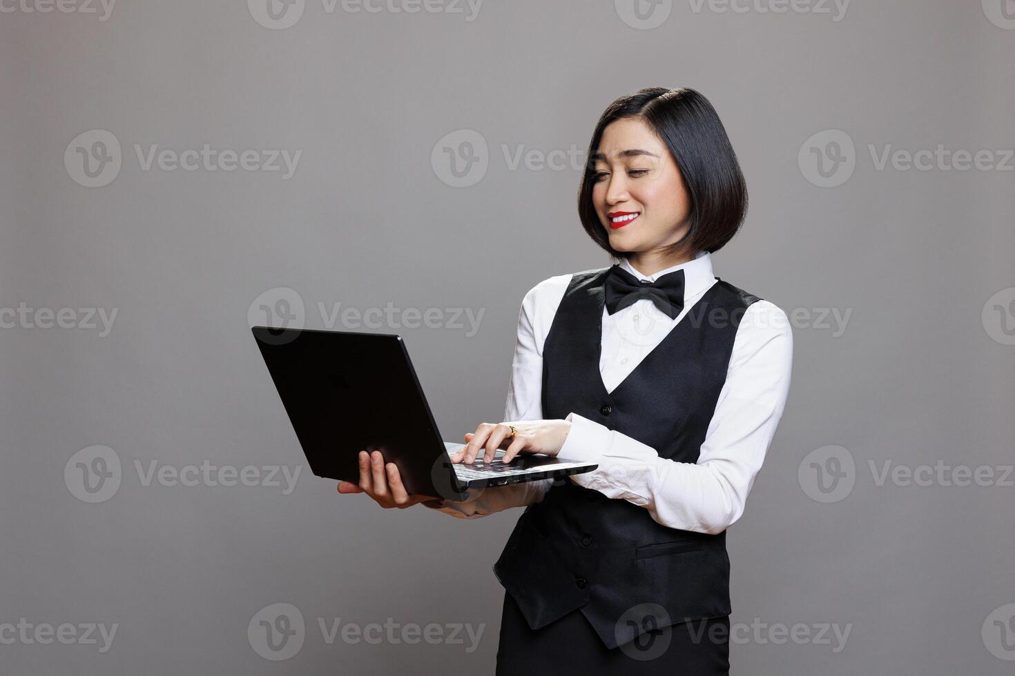 Smiling asian waitress in professional uniform using laptop, checking catering service orders. Cheerful woman receptionist holding portable computer and typing on keyboard photo
