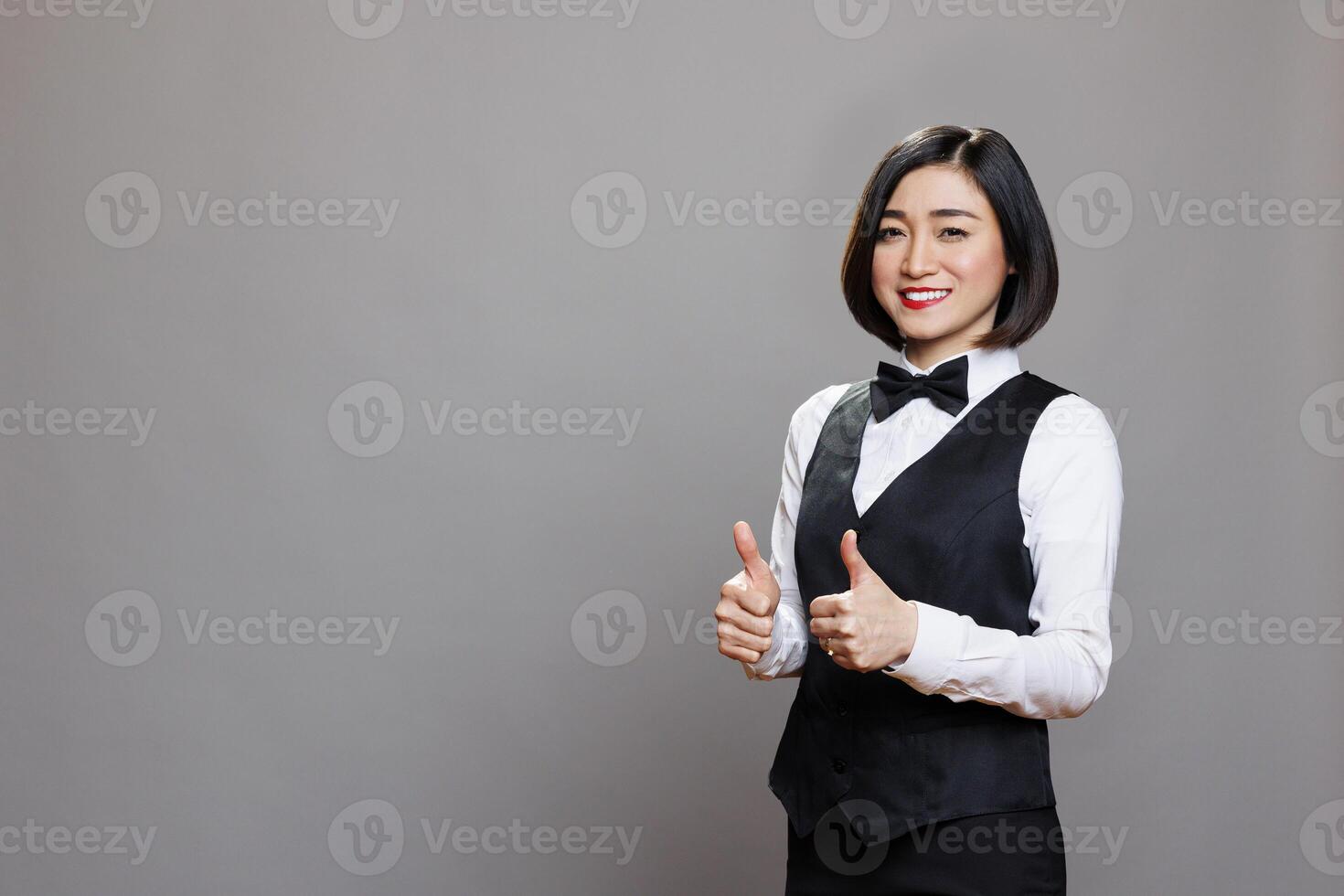 Cheerful woman receptionist showing two thumbs up symbol studio portrait. Smiling asian waitress standing with like gesture and looking at camera with happy face expression photo