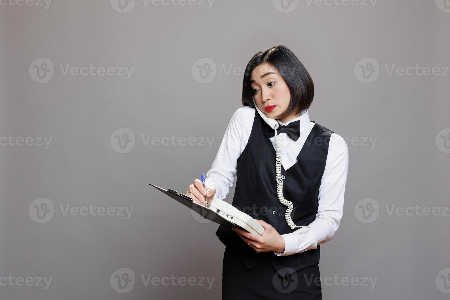 Confused young asian waitress managing conversation on landline phone and writing on clipboard. Restaurant receptionist in uniform maintaining records while chatting with customer on telephone photo