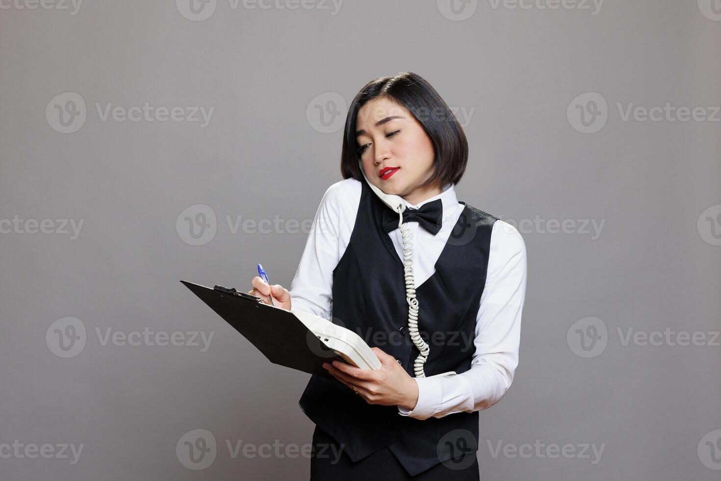 Young asian waitress dressed in uniform writing on a clipboard while talking on landline phone. Restaurant young receptionist chatting with manager on telephone and taking notes photo
