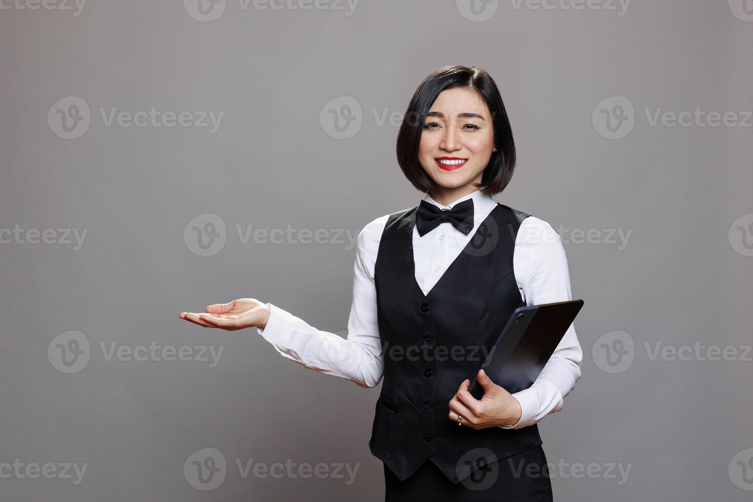 Smiling asian waitress holding digital tablet and welcoming guest with hand portrait. Restaurant receptionist standing with portable device and showing with arm while looking at camera photo