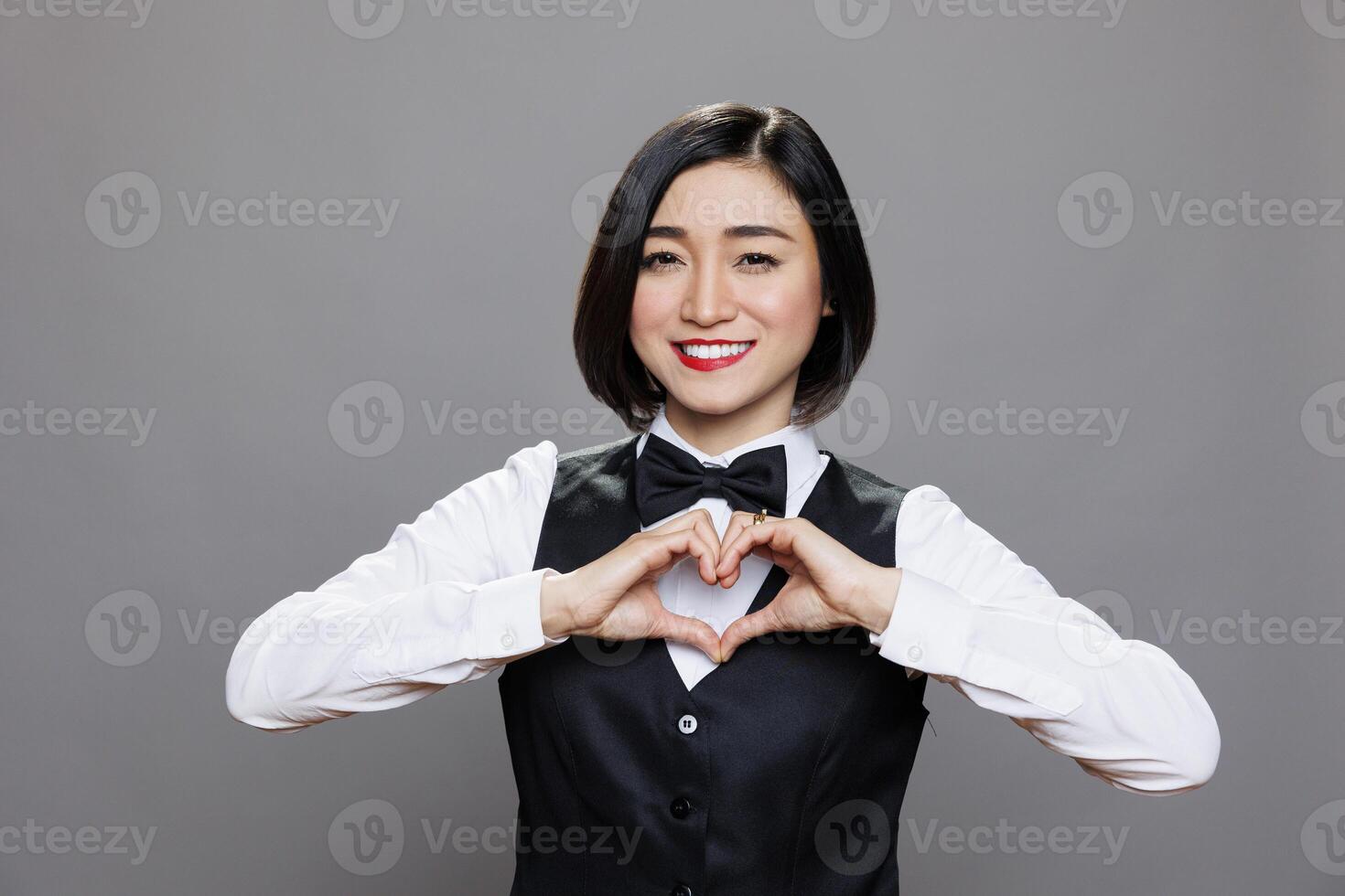 Smiling asian waitress showing heart shape with hands, sharing love portrait. Cheerful woman receptionist showcasing affection symbol and looking at camera with positive face expression photo