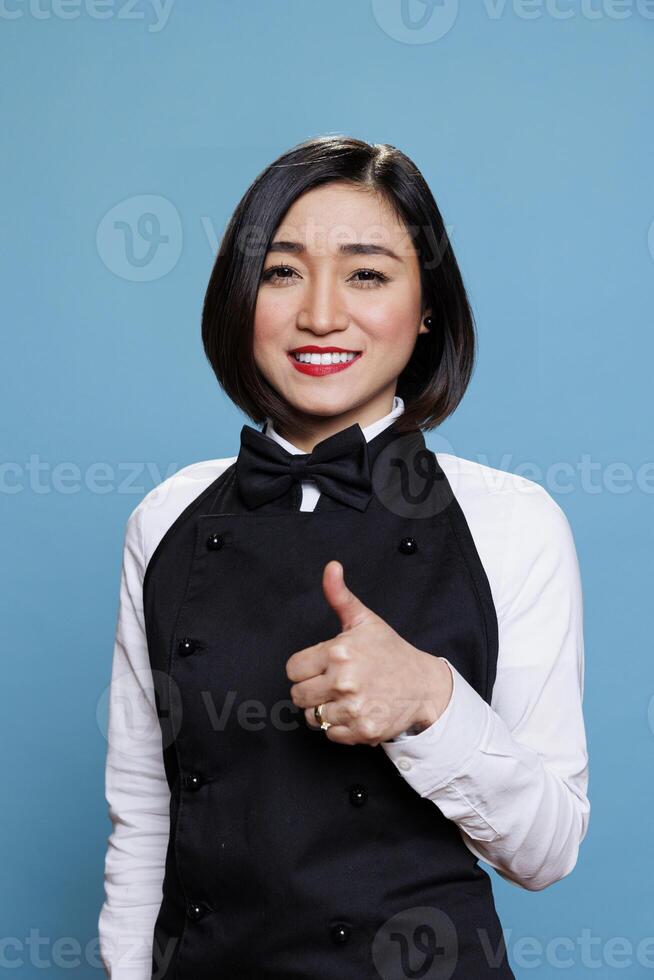 Asian waitress displaying satisfaction with thumb up gesture and looking at camera with cheerful face. Joyful woman receptionist expressing approval with like symbol portrait photo