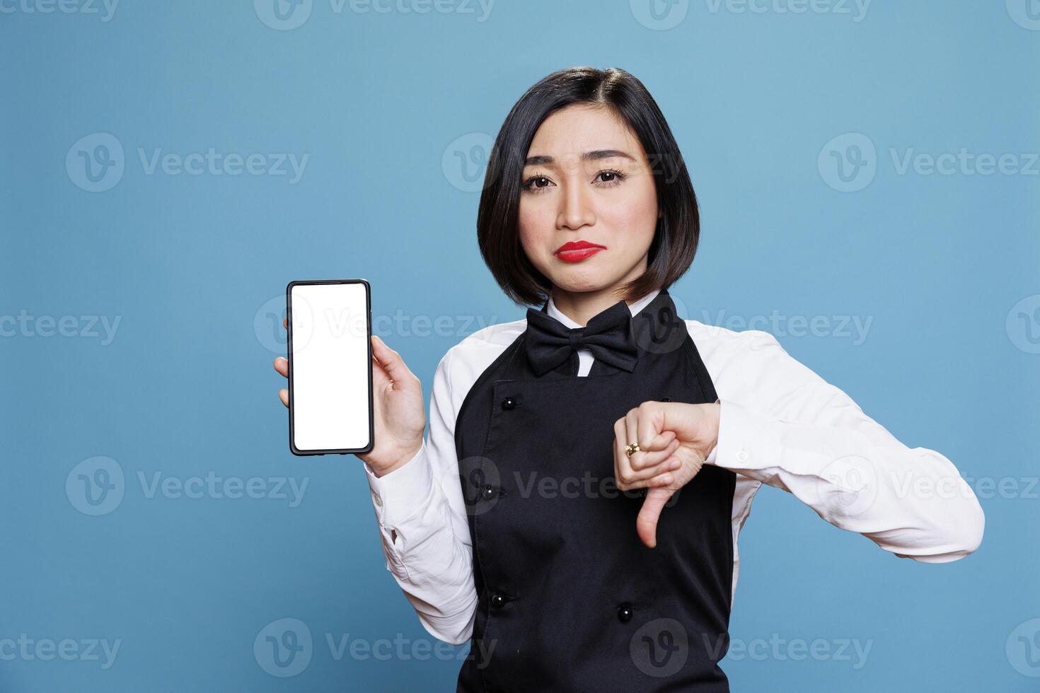 Asian waitress displaying smartphone blank touchscreen mockup and showing thumb down gesture portrait. Disappointed woman receptionist presenting mobile phone empty screen and expressing dissatisfaction photo