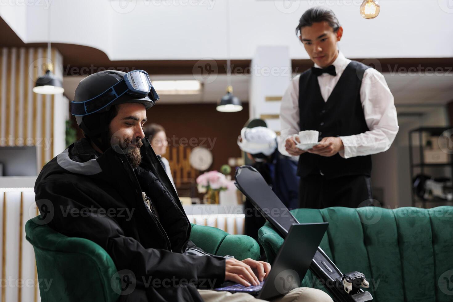 Man dressed in winter jacket using laptop in lounge area while asian waiter brings beverage. Male tourist booking activities with digital device for his skiing vacation at luxury winter resort. photo