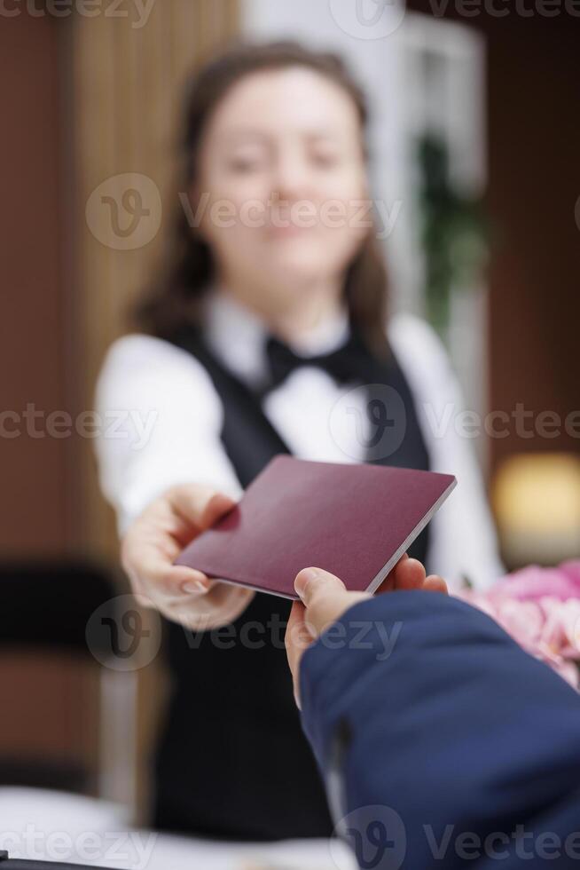 Close-up shot of female concierge returning identity card to traveler wearing winter jacket after check-in process at front desk. Detailed view of passport being grasped by receptionist and tourist. photo