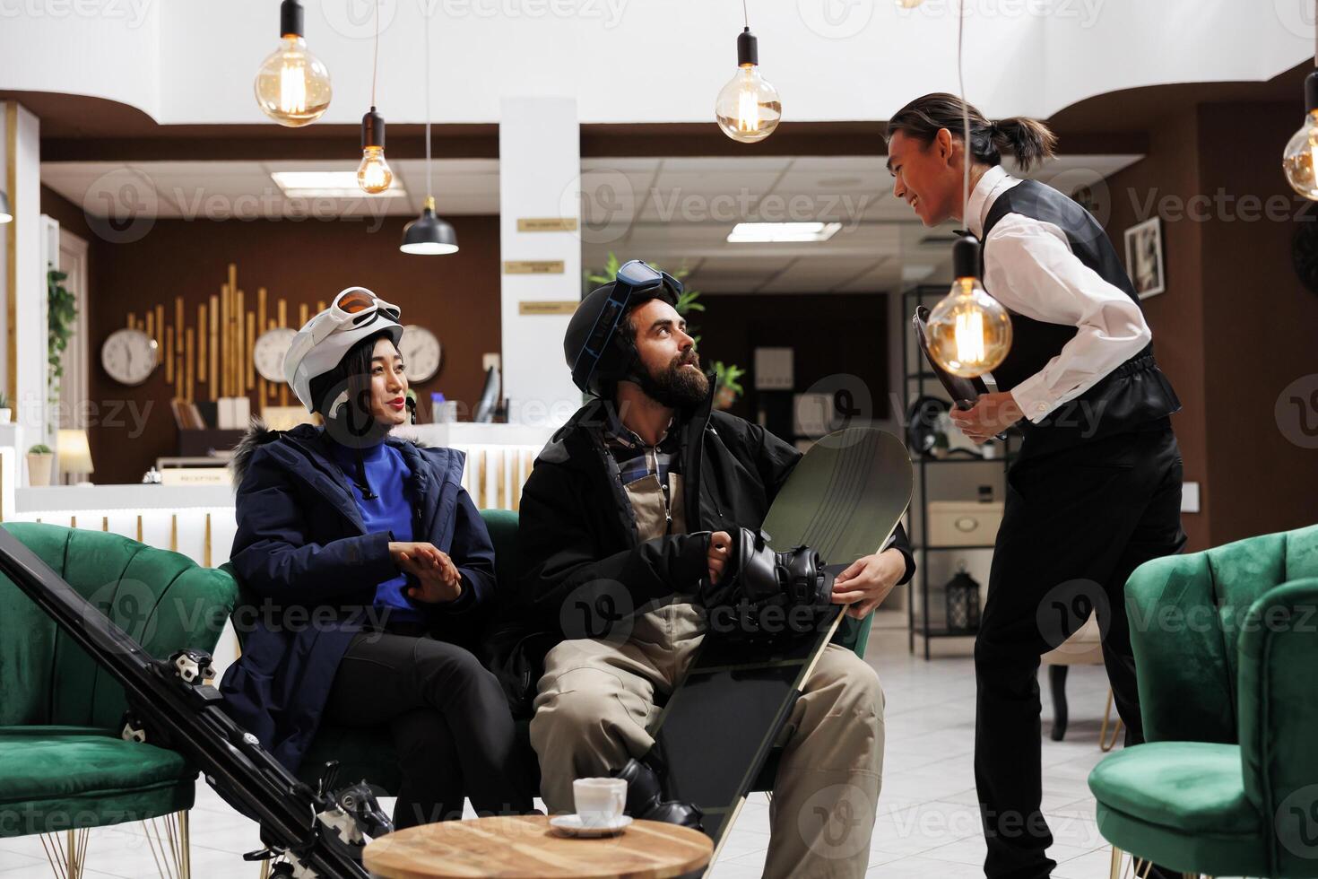 Excited couple with skiing and snowboarding gear receive assistance from friendly employee in hotel lobby. Visitors embrace winter adventure at ski resort ready for holiday activities. photo