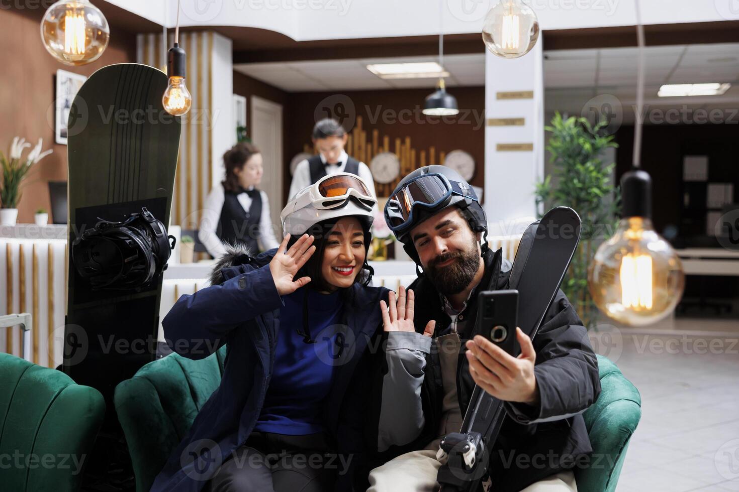 Tourists enjoying winter vacation at ski resort dressed in snow gear. Young couple use mobile device to capture the moment while surrounded by employees and wintersport equipment in hotel lobby. photo
