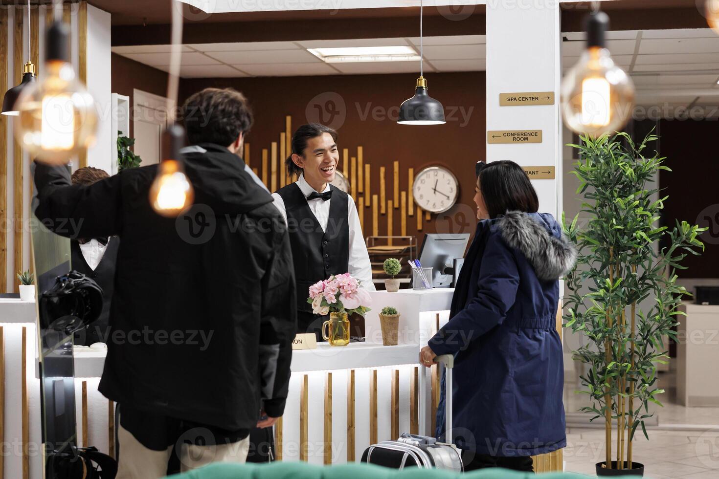 Young couple with winter clothes and suitcase arriving at front desk of luxury holiday resort for room reservation. Friendly concierge welcomes two tourists with ski attire at hotel reception counter. photo