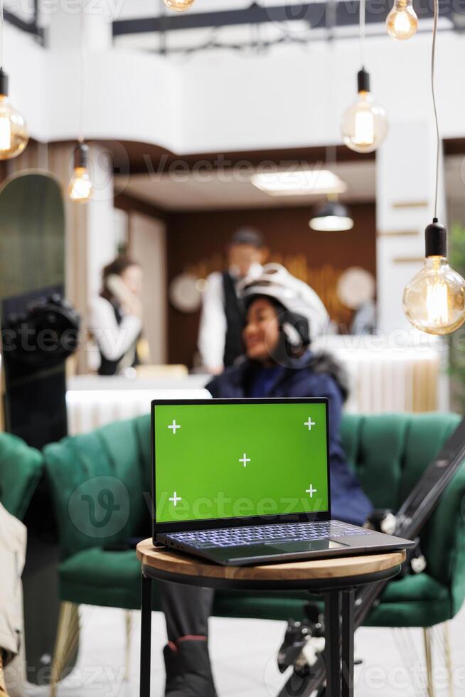 Detailed shot of digital device with isolated copyspace template placed on bench in lounge area of luxury ski mountain resort. Woman in wintersports gear at background of laptop with green screen. photo