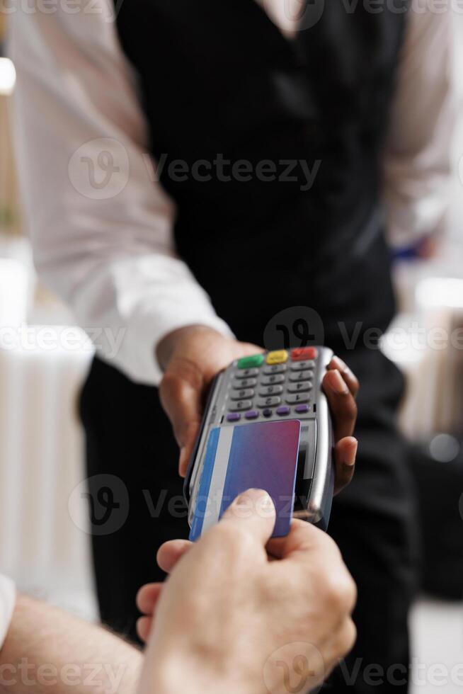 Photo focus of elderly caucasian man tapping his credit card on payment machine held by waiter in lounge area. Detailed view showing a person paying on pos terminal for reservation in hotel lobby.