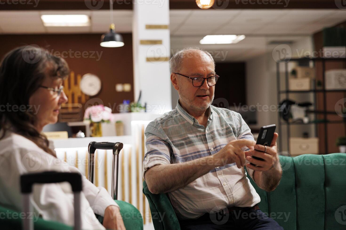 On couch in lounge, senior guy visitor using smartphone to browse the internet. Retired elderly husband utilizing mobile device to make online reservations while sitting on comfortable sofa with wife. photo