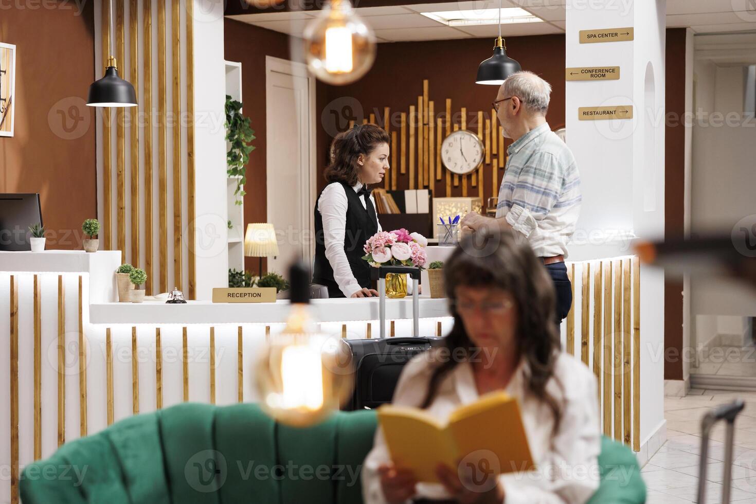 Retired senior man checks in at front desk with help from receptionist in hotel foyer. Elderly woman with book, relaxing in comfortable lounge area while male tourist waits at counter. Vacation vibes. photo