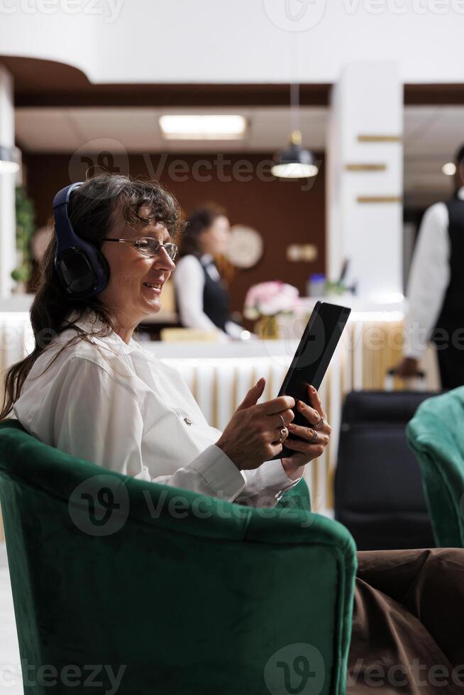 Side-view of smiling eldery lady with wireless headset, browsing on tablet for holiday activities. In hotel lobby, old woman in close up shot, wearing headphones and surfing the net on digital device. photo