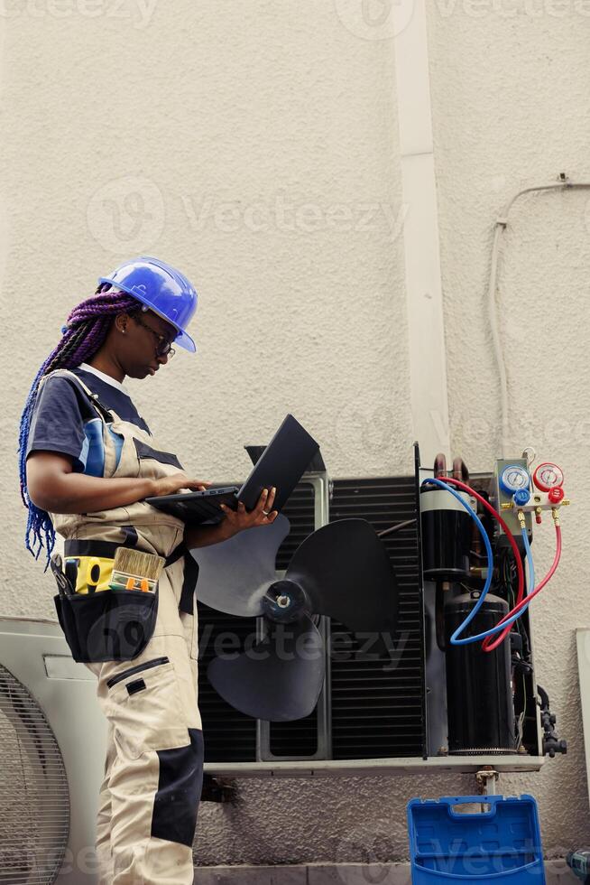 Licensed technician commissioned for outside hvac system annual maintenance, looking for refrigerant leaks. Trained engineer doing air conditioner inspection, adding data on laptop photo