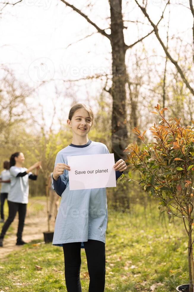 Portrait of young kid advertising banner with save our planet message against pollution and illegal dumping, volunteering to preserve forest habitat. Small child holds awareness paper sign. photo