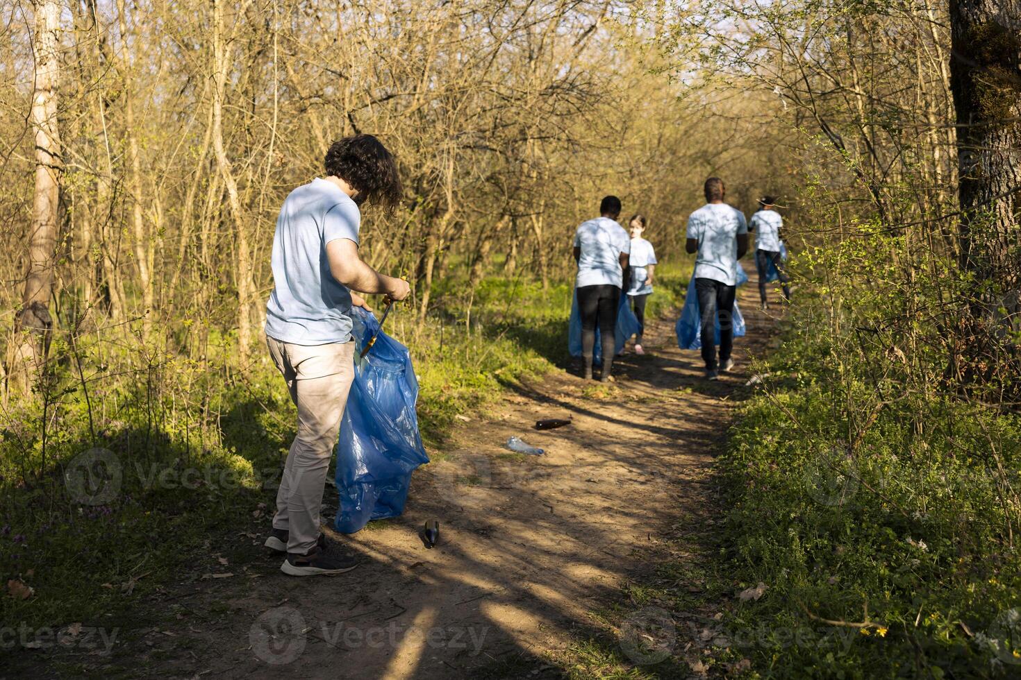 Male environmentalist collecting trash with a long claw tool and bag, grabbing plastic waste and rubbish to tidy up the forest area. Young man cleaning the natural habitat, global pollution. photo