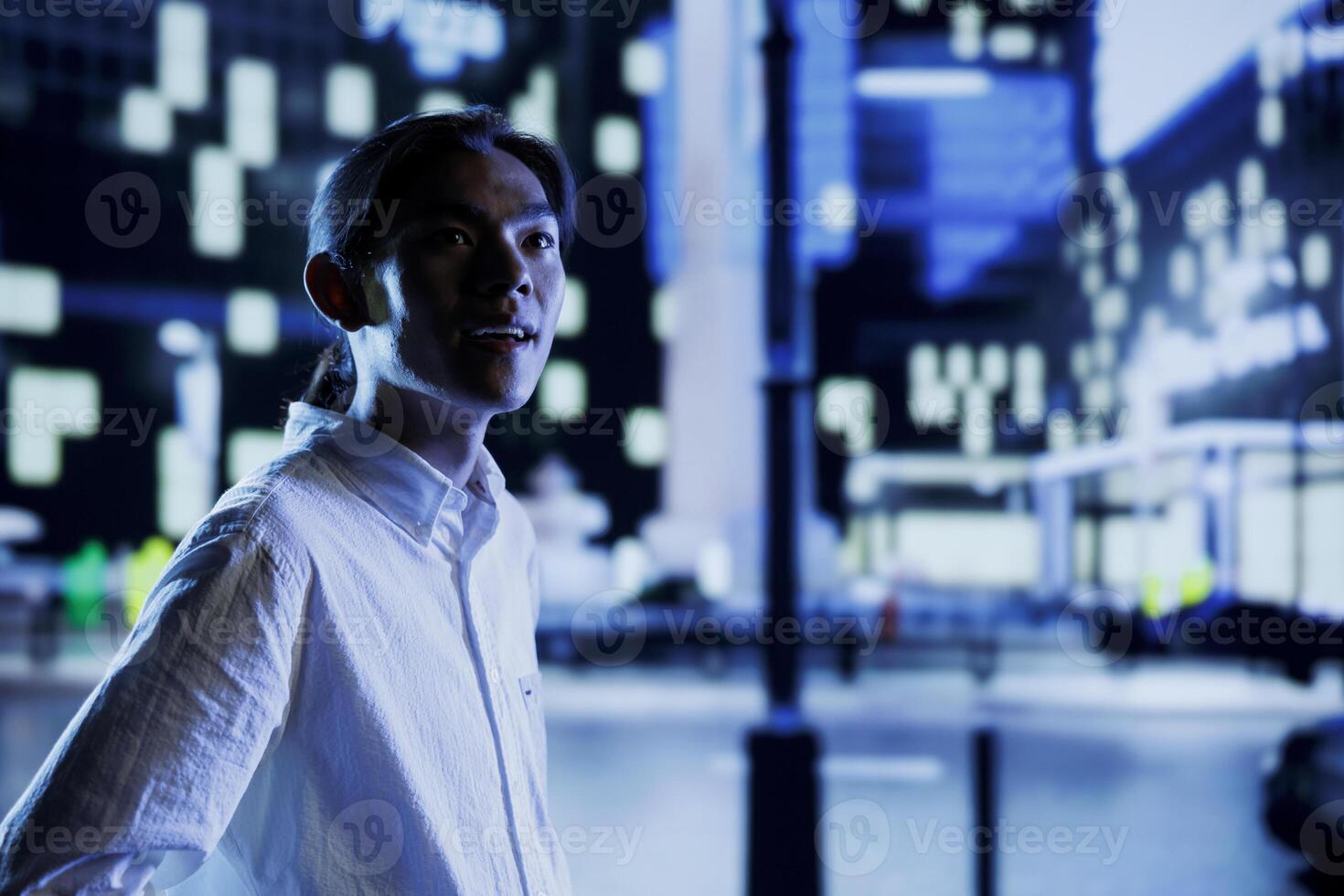 Asian man wandering around city boulevards during nighttime, being amazed by nightlife environment buildings. Citizen strolling around empty avenues at night, enjoying metropolitan skyscrapers photo
