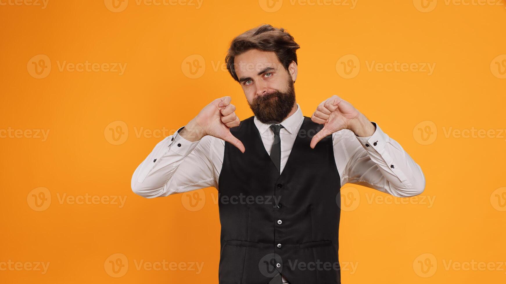 Staff member gives thumbs down on camera, showing negative dislike sign against yellow background. Man restaurant employee expressing disagreement with bad gesture, displeased butler. photo