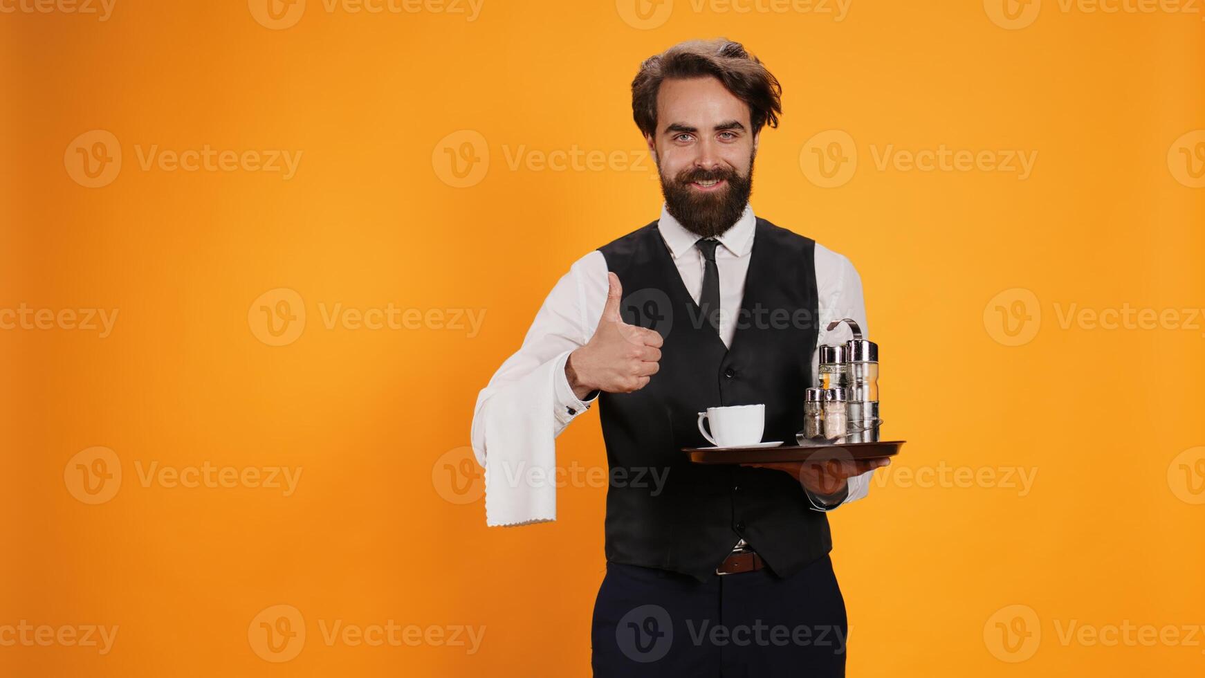 Anccomplished valet raises thumbs up in studio, displays agreement emblem, is enthusiastic on camera. Before serving beverage, young man with a waiter job gestures approbation with like sign. photo