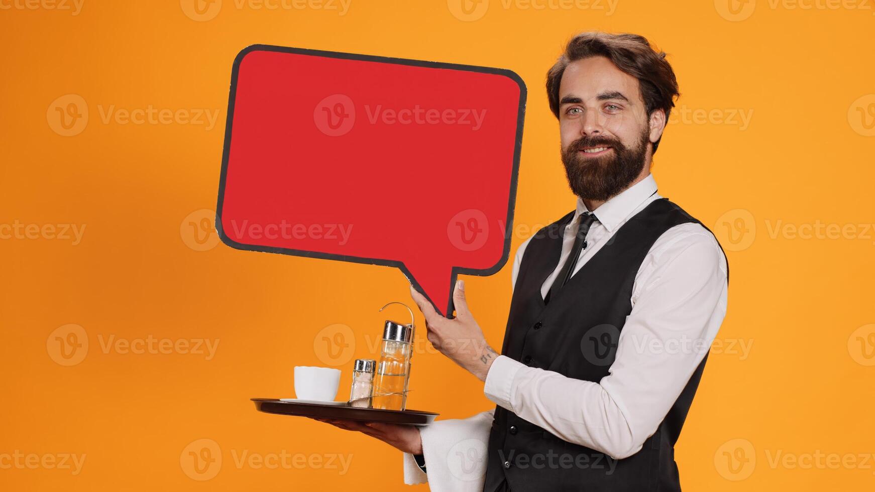 Classy waiter holds speech bubble icon on camera, working to create new advertisement using empty red cardboard. Experienced employee in suit presenting blank carton billboard, serving personnel. photo