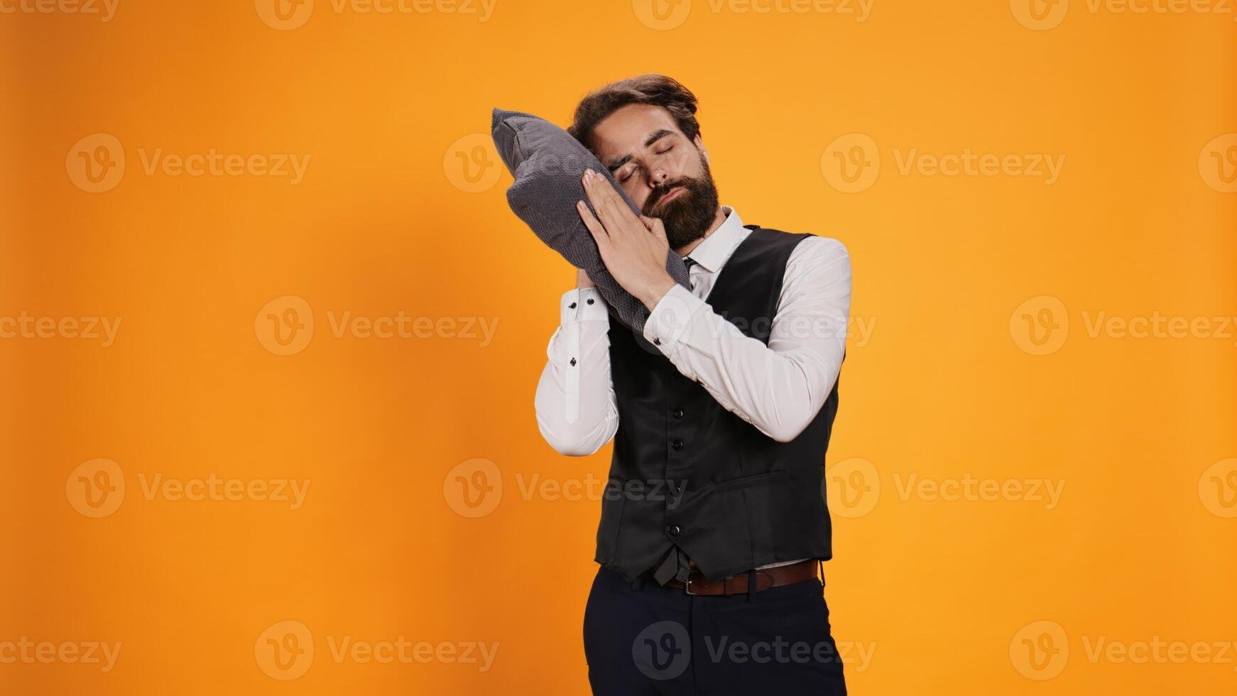 Tired diner employee napping on pillow, sleeping while he in standing in front of camera. Overworked waiter falling asleep against yellow background, experiencing burnout in restaurant sector. photo