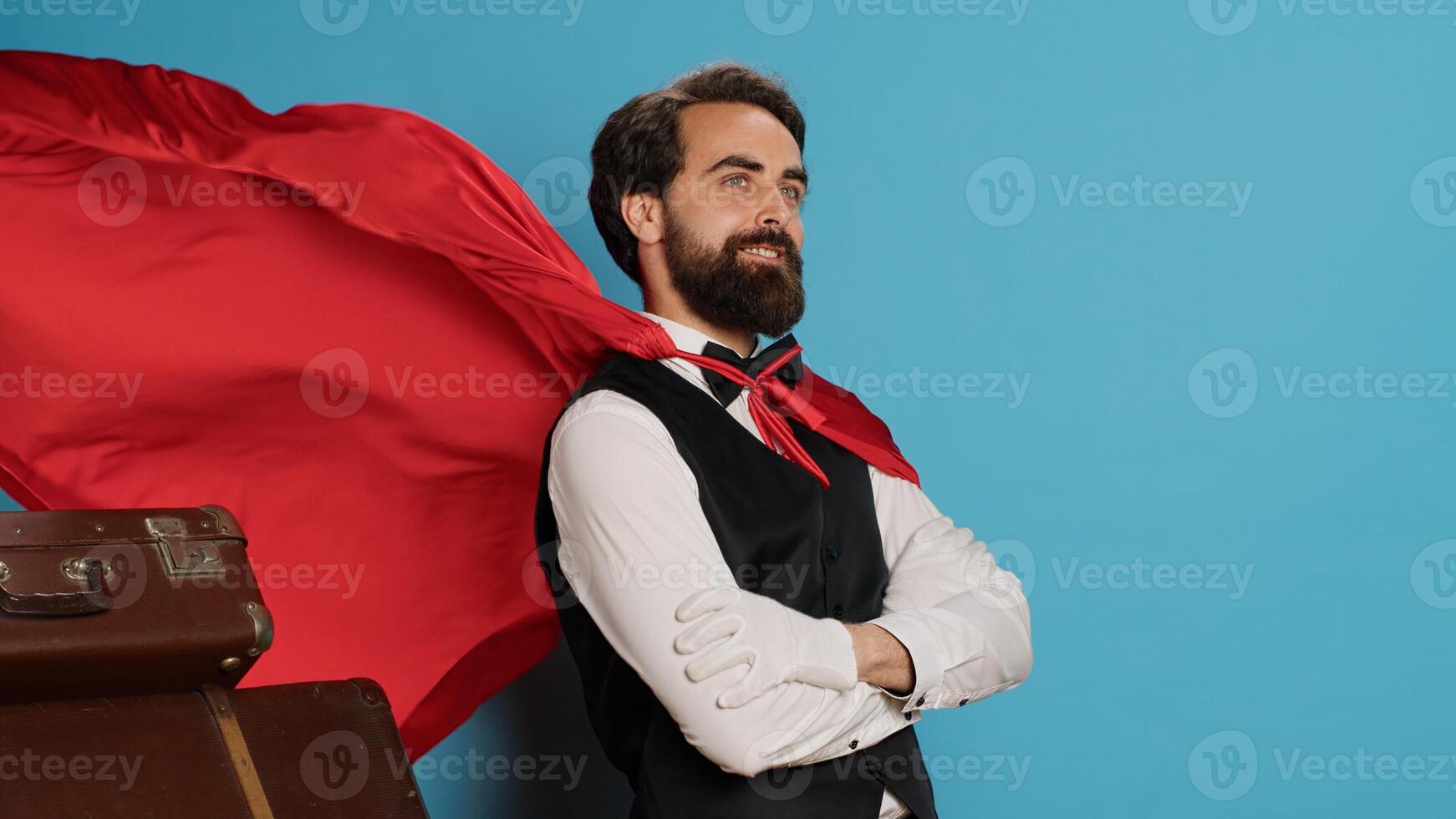 Hotel concierge with red hero cape to help guests with baggage, pretending to be cartoon superhero character on camera. Bellhop acting like fantasy mascot with mantle next to trolley bags. photo
