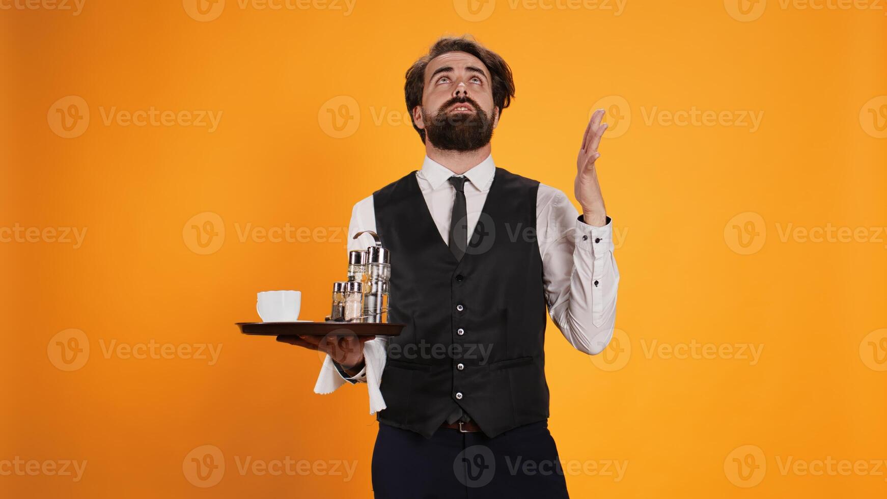 Happy man acting spiritual worshiping Jesus in studio and seeking fulfillment. Working at posh restaurant with platter, devoted religious waiter looks delighted against yellow background. photo
