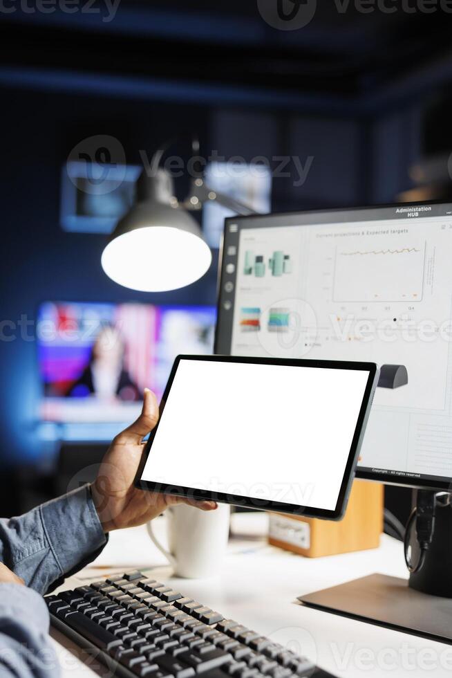 Young employee checks tablet with white screen at home, calculating finances and sales trend on computer. Advisor uses pc and device with isolated empty layout, monthly statistics. photo
