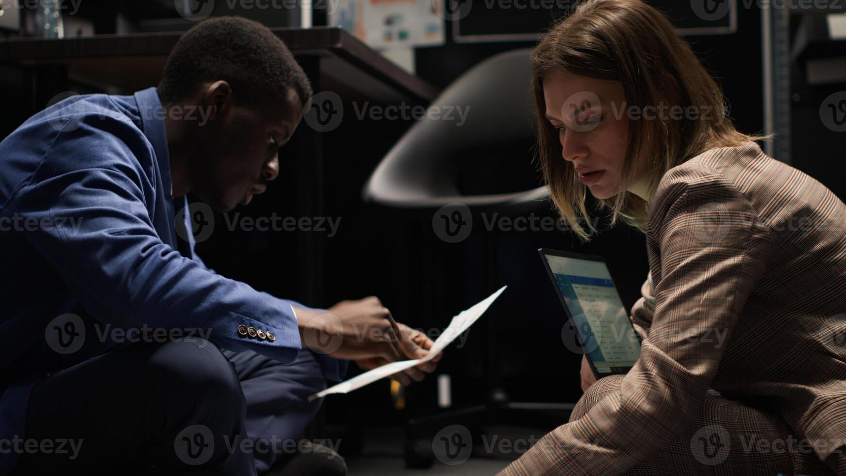 Multiethnic private investigators pursuing the truth in crime case, gathering evidence in incident room. Black policeman and caucasian detective sitting on floor surrounded by clues, files and laptop. photo