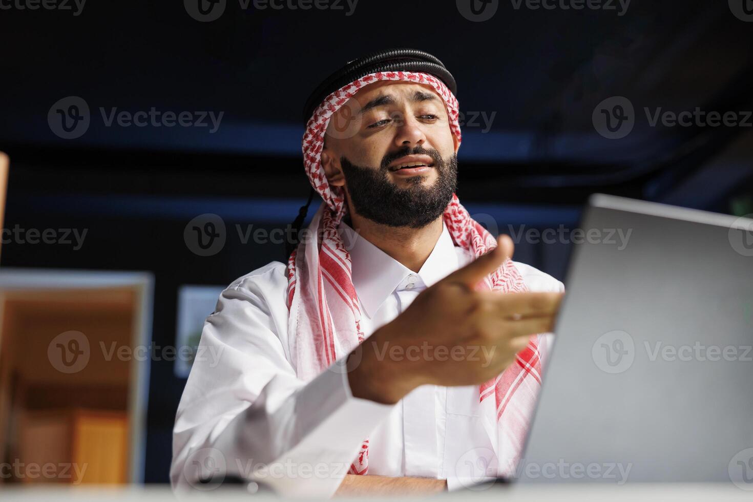 Young businessman representing success in the digital era, wearing Arabian apparel, using a digital laptop. Muslim guy conversing confidently on his personal computer at his home office desk. photo