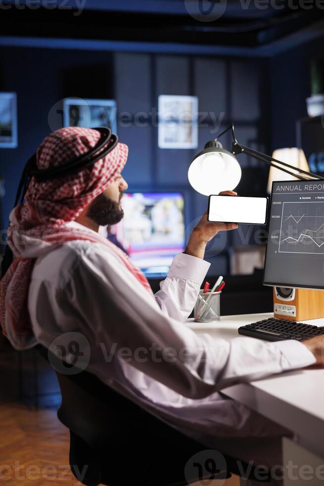 Side-view shot Middle Eastern man in traditional attire sitting at an office desk, using a desktop computer. They hold a mobile phone with a blank chromakey mockup template, offering space for advertising. photo