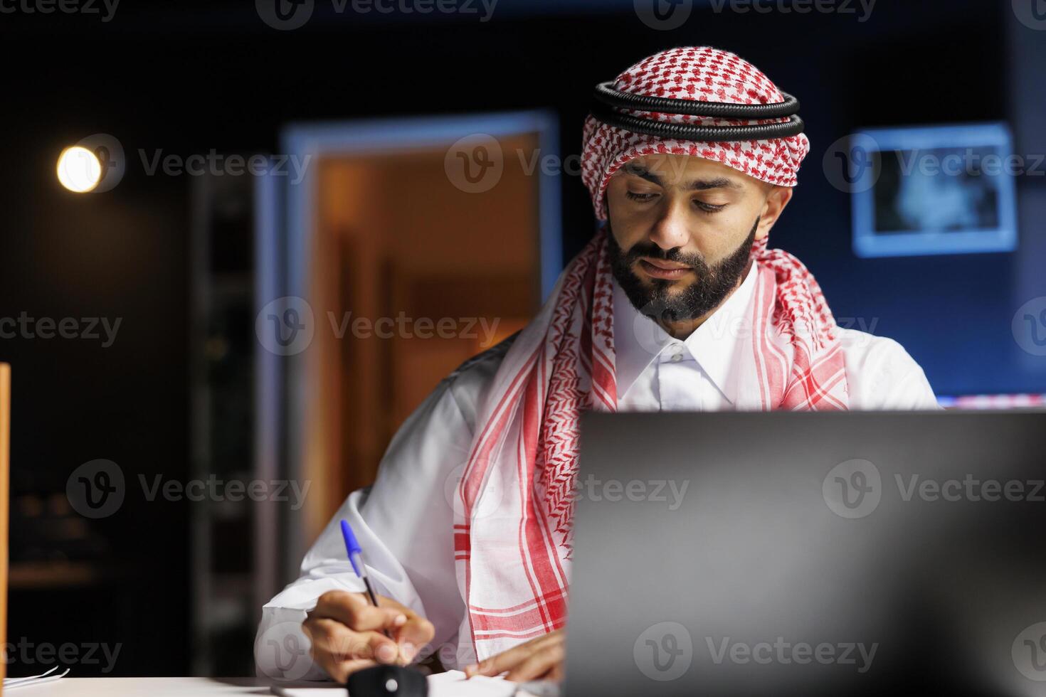 Male Arab entrepreneur working at his modern desk, browsing the internet and writing in his notebook. Image shows a young Muslim man using a pen, book and a minicomputer on the table. photo
