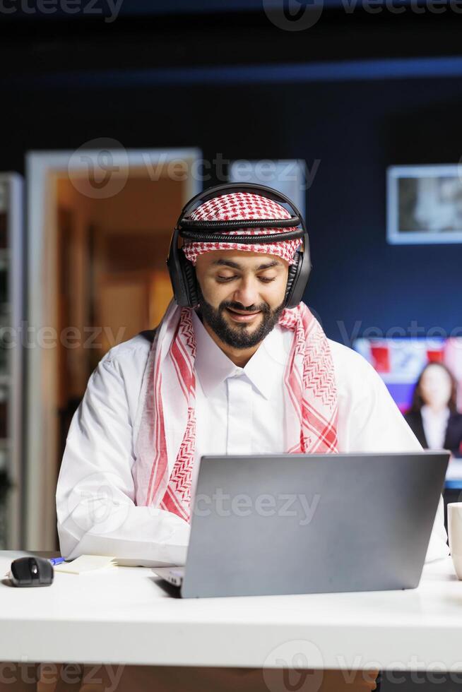 Professional Arab guy at his desk, diligently using his laptop for research. He engages in conferencing and email communication, showcasing a blend of traditional attire and wireless technology. photo