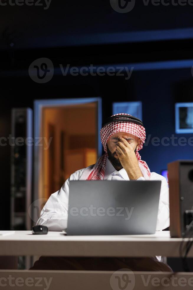 Exhausted businessman dressed in Arabic clothing sits at desk trying to conduct internet research on personal computer. Male Muslim person with a laptop on the table seems worn out and has a headache. photo