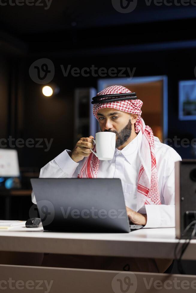 Youthful Muslim businessman with a cup, working on his minicomputer, indoors at a desk. At the table, an Arab male efficiently uses a digital laptop while drinking coffee. photo