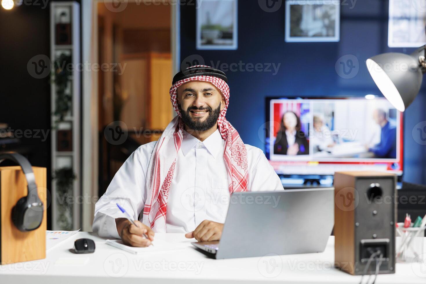 Middle Eastern male freelancer in traditional attire, browsing the internet on laptop while holding a pen. Arab man smiles confidently at camera, reflecting his professional profile in modern office. photo