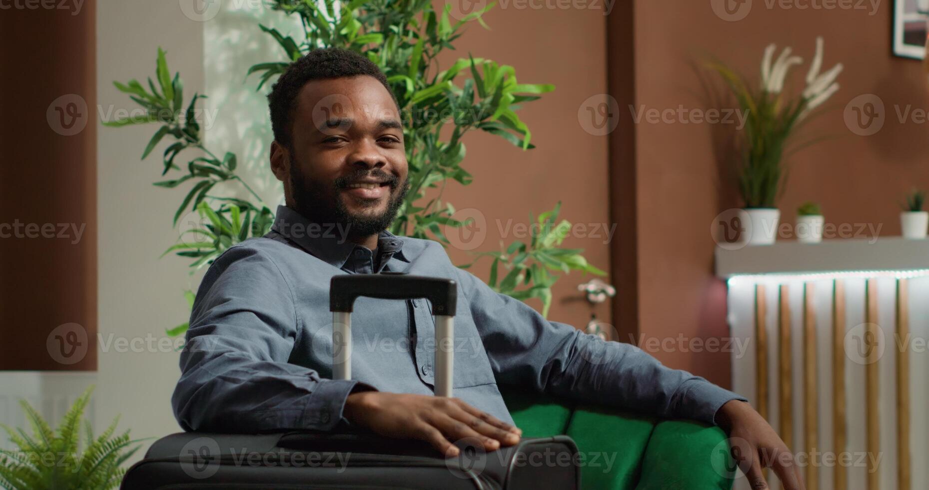 African american guest preparing for check in, relaxing in comfy chair while holding luggage, ready to sign for accommodation during trip. Cheerful person relaxes in lobby lounge area. Handheld shot. photo