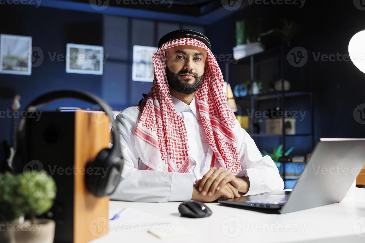Portrait shot of an Arab businessman in traditional attire sitting at a desk with his personal computer. The image shows a Muslim guy looking at the camera in his professional office. photo