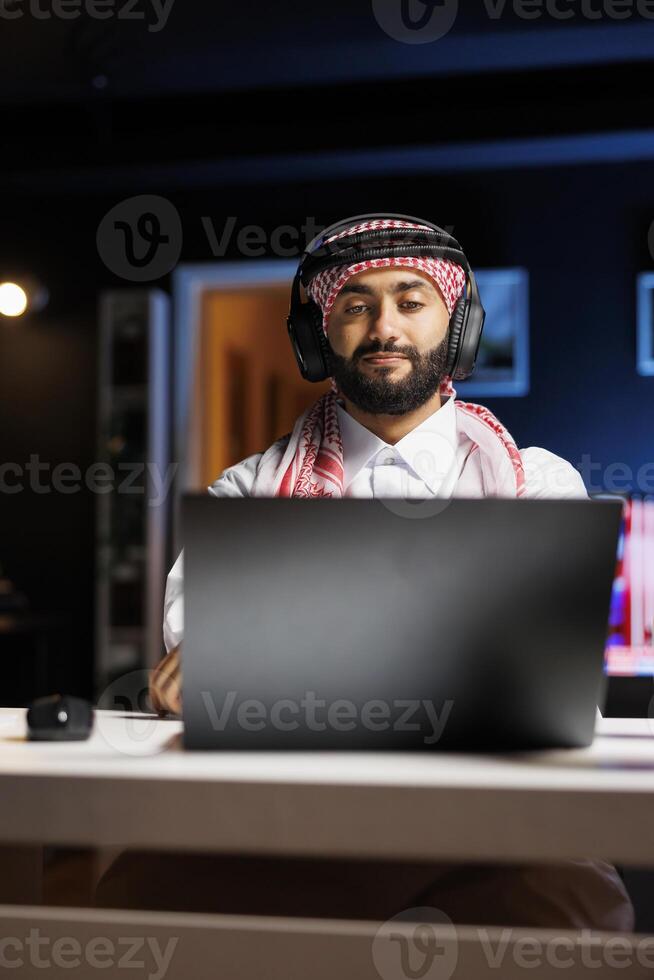Middle Eastern man in traditional attire efficiently uses a laptop for research and communication via email. Arab guy with wireless headphones having a call on his personal computer. photo