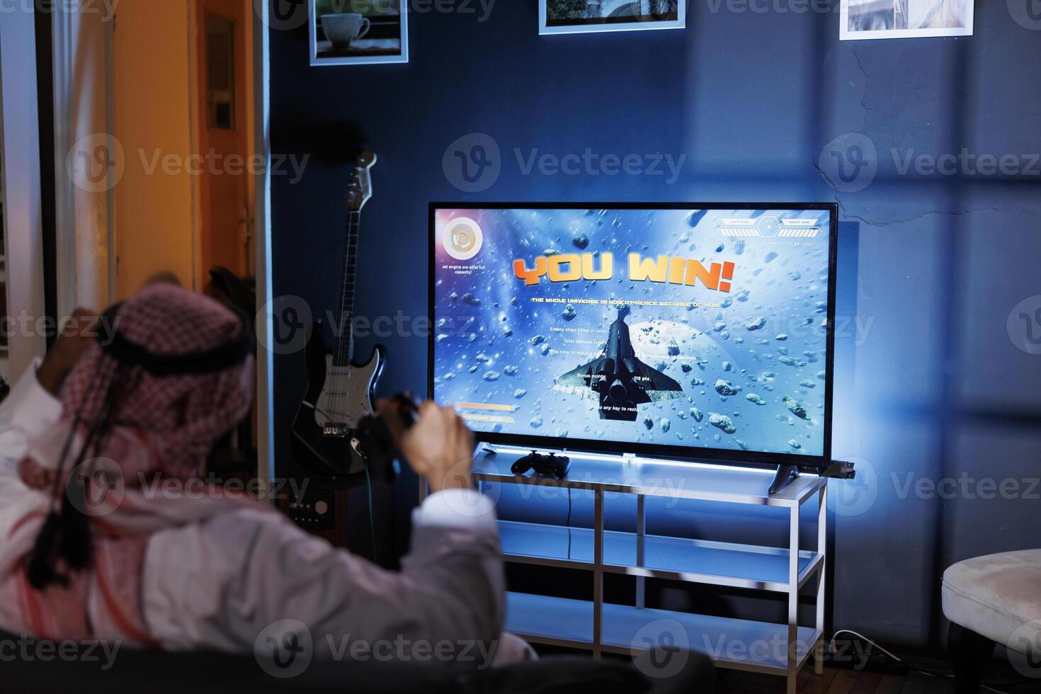 A young Arab man plays an immersive virtual reality game at home, celebrating victory while holding a wireless controller. He is sat at home, absorbed in amusement, and lighted by the screen light. photo