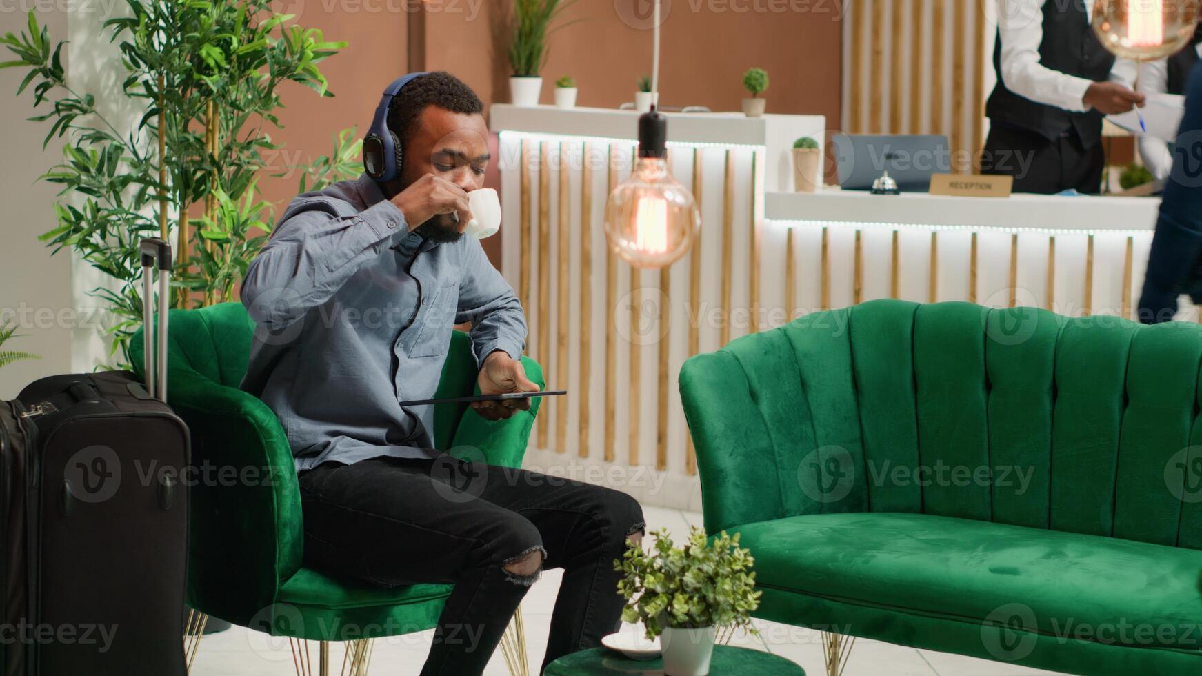 Visitor listens to music at reception, waiting to do check in registration at front desk in lobby. African american guest using headset in lounge area, having fun with songs after travelling. photo
