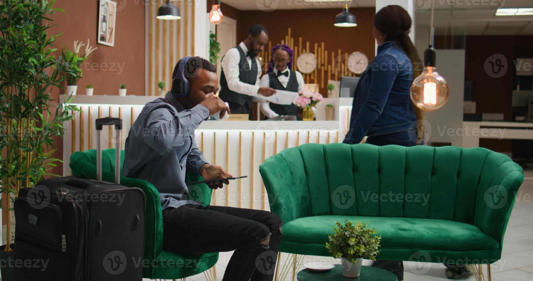 Client listens to music at hotel reception, preparing to sign registration papers waiting to do check in at front desk in lobby. African american person using headset in lounge area, tourism. photo
