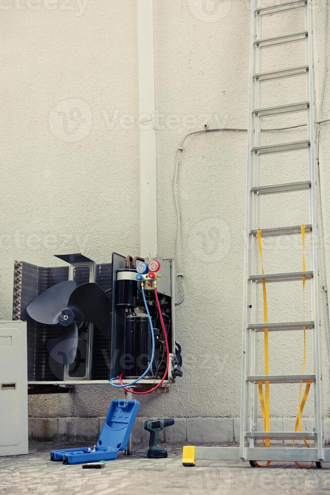 Malfunctioning external hvac system unit in need of reconditioning next to technician working gear. Outdoor opened air conditioner and professional folding ladder, toolkit, power drill photo