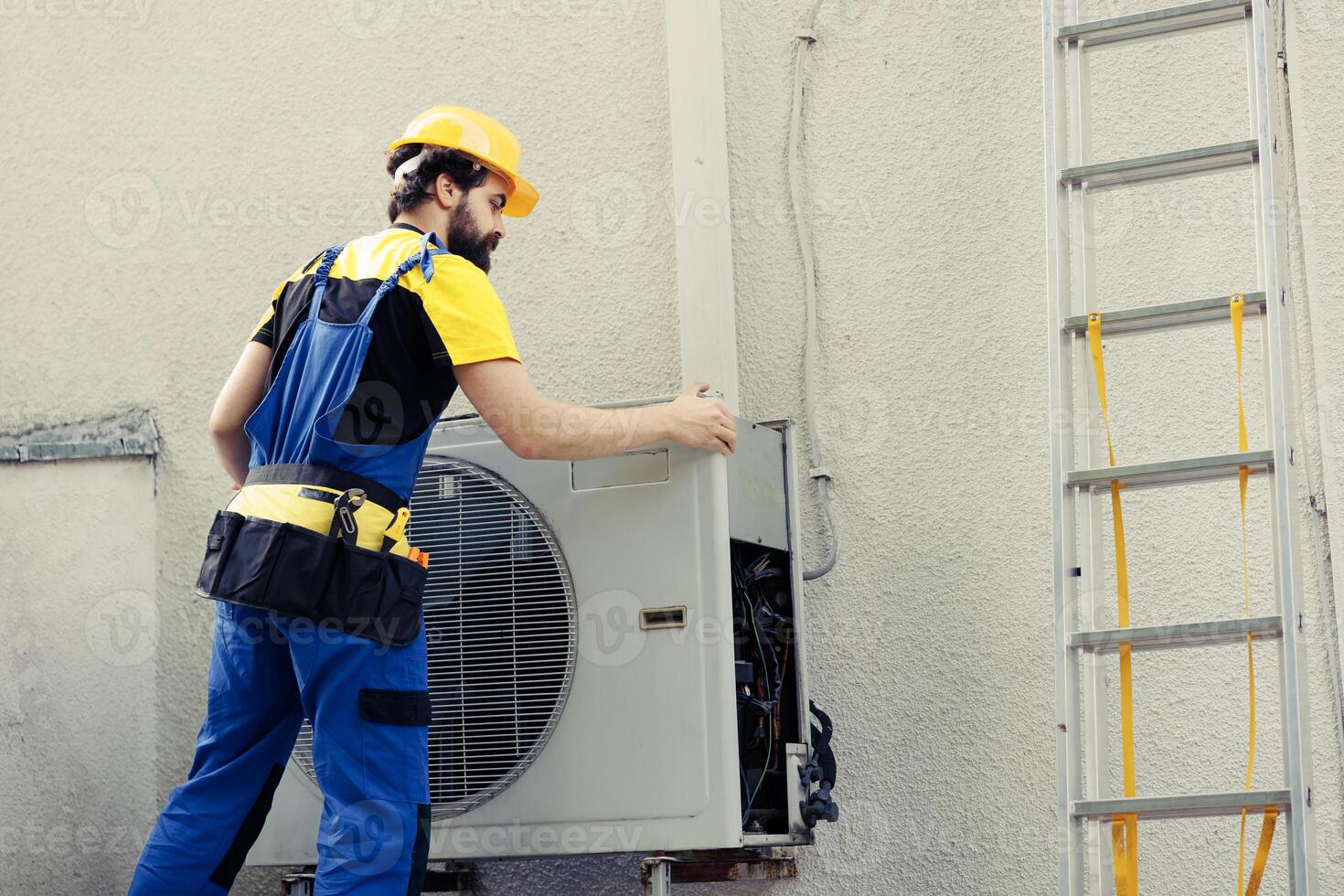 Skilled wireman starting work on malfunctioning air conditioner, using electric drill to take apart condenser coil panel. Experienced serviceman dismantling hvac system to check for faulty wires photo