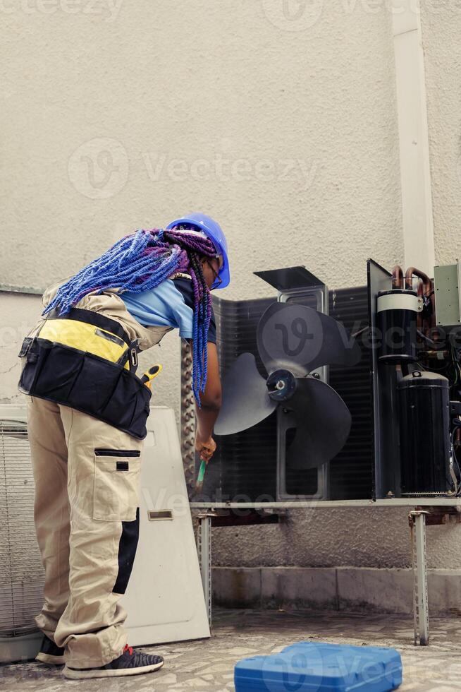 African american skilled mechanic commissioned by home owner for annual air conditioner routine maintenance, using soft dusting brush to clean dusty condenser coils and filters, outdoor photo